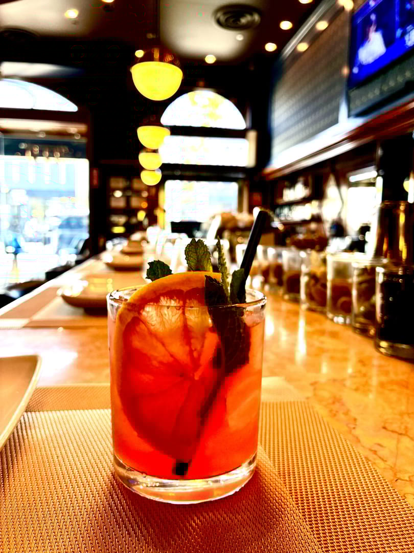 A vibrant red cocktail with orange slices and mint sits on a bar counter with warm lighting.